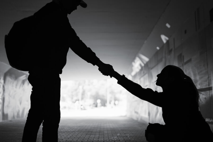 Silhouette of a man and a woman in a dark urban tunnel. The man is wearing a backpack and baseball hat and is standing. The woman is in a jacket and has a ponytail. She is kneeling on the bricked walkway, looking up at him, as she accepts his helping hand.